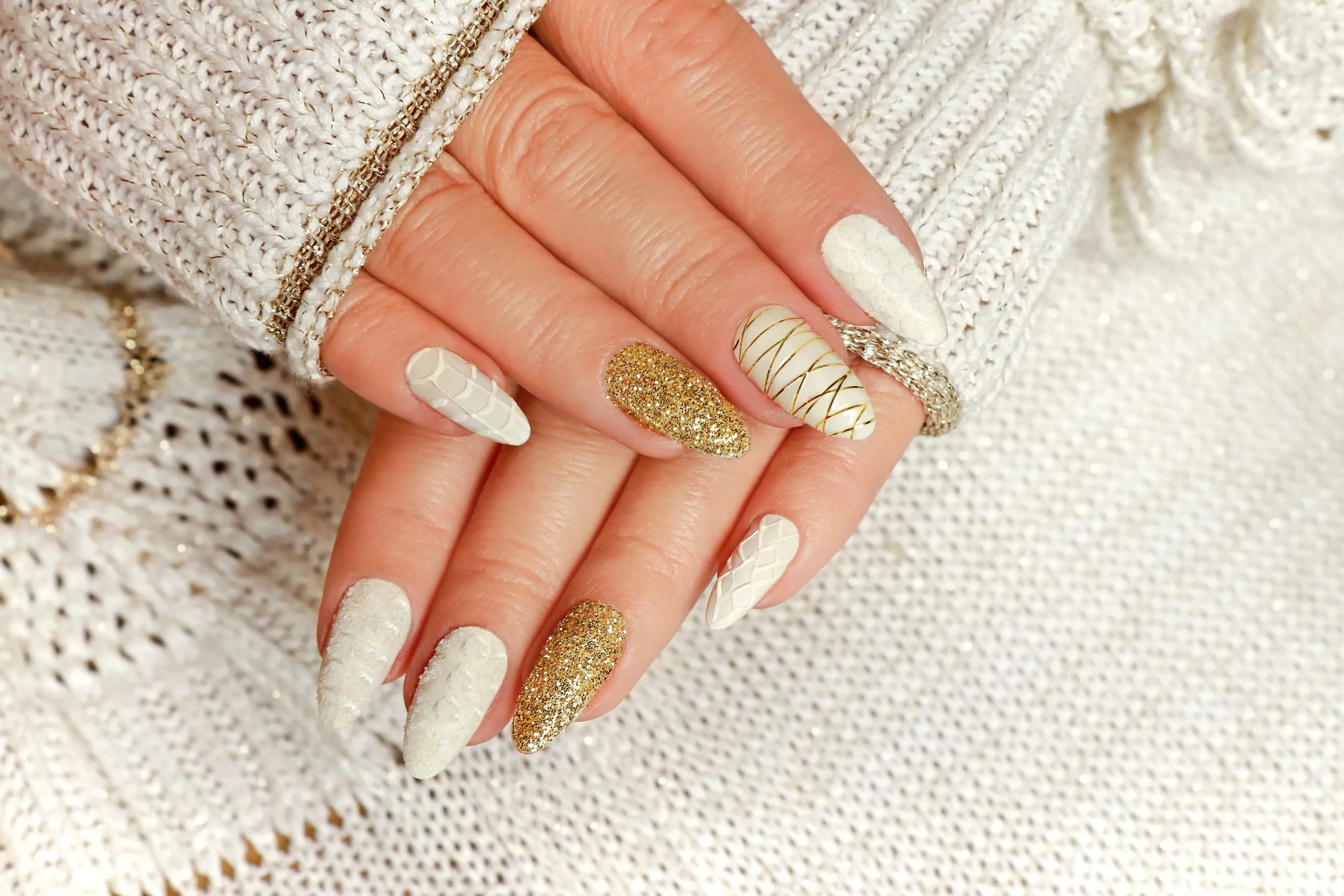 Manucure longue en amande avec motifs blancs texturés et paillettes dorées.